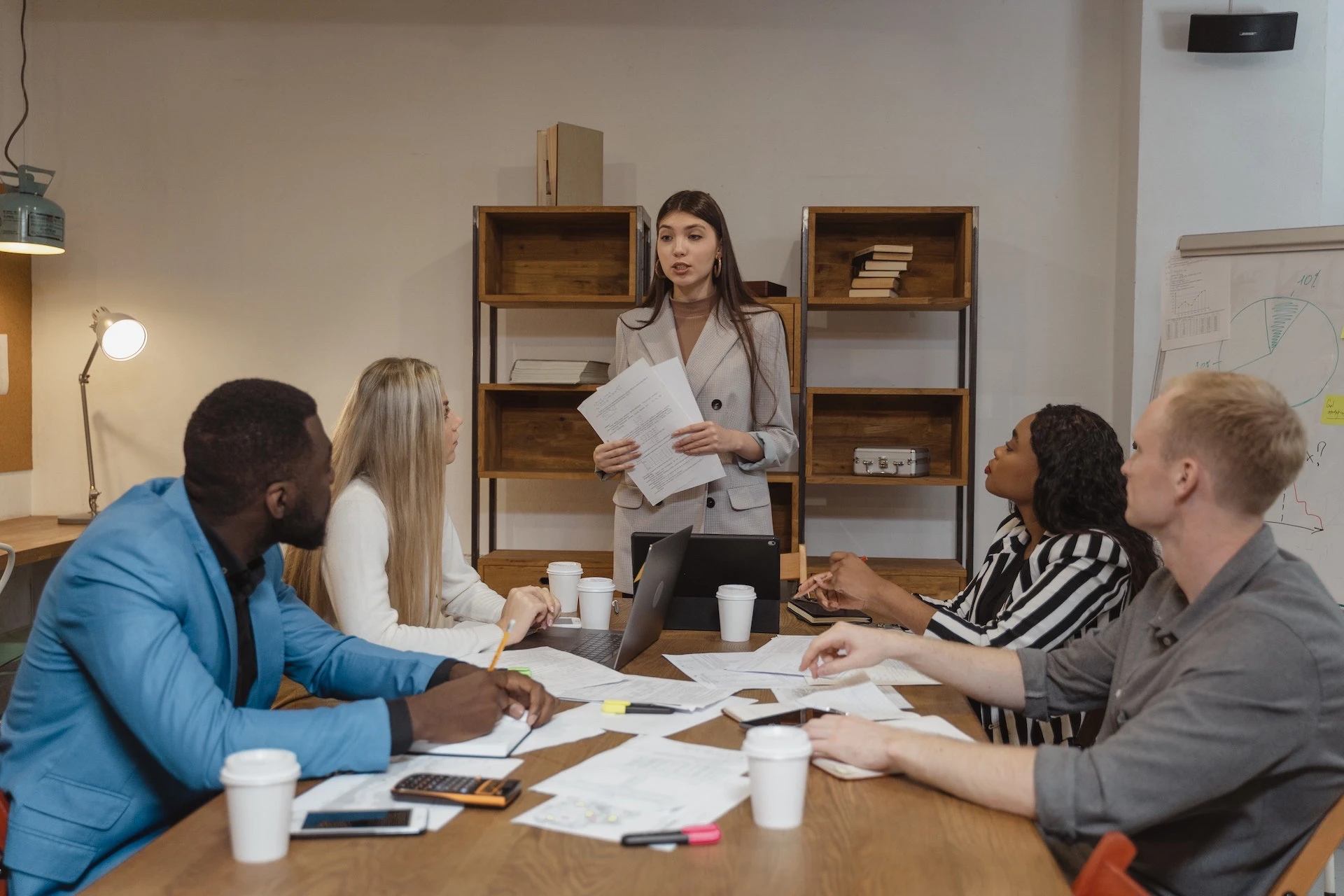 eine Führungskraft steht vor einem Team aus vier Personen, die gerade an einem Tisch sitzen und arbeiten, und spricht zu ihnen.